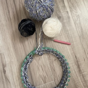 May include: A craft image featuring a circular knitting loom with blue and white yarn wrapped around it. Three balls of yarn in blue, black, and white are positioned above the loom. A pink crochet hook rests nearby, suggesting a knitting project.