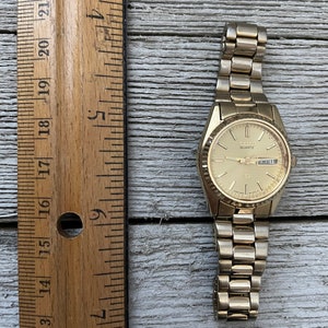 May include: A gold-toned Seiko Quartz wristwatch with a gold-tone metal band. The watch face is white with black markings and a date window. The watch is about 2.5 inches in diameter.