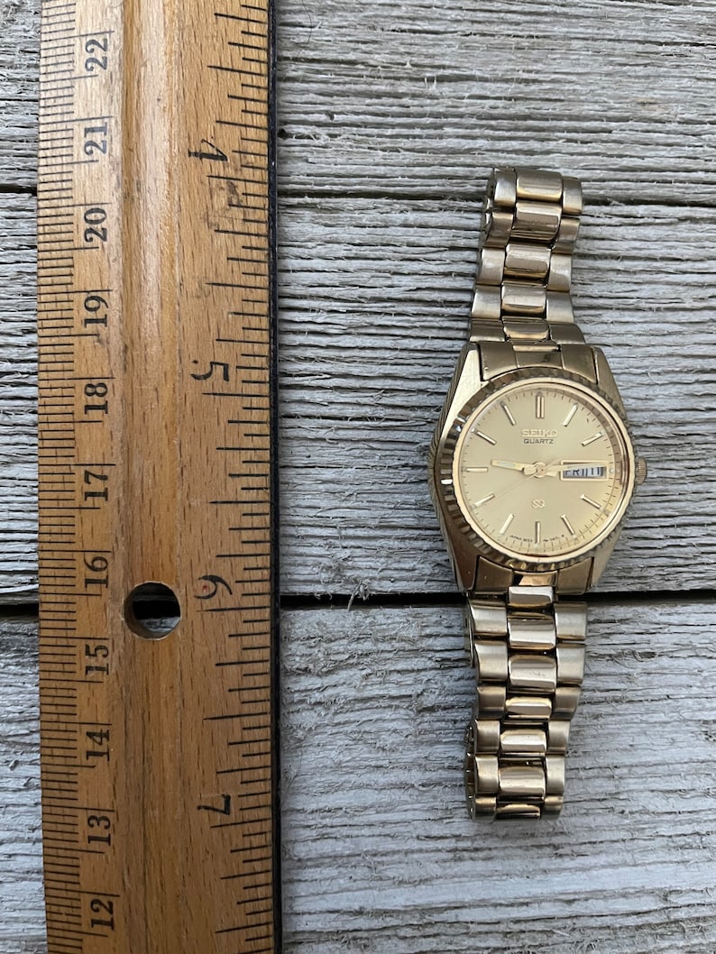 May include: A gold-toned Seiko Quartz wristwatch with a gold-tone metal band. The watch face is white with black markings and a date window. The watch is about 2.5 inches in diameter.
