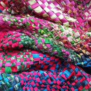 May include: A woven tapestry made from strips of fabric in shades of pink, green, blue, and black. The fabric is woven in a checkered pattern.