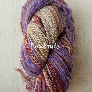 Puede incluir: Un ovillo de hilo en tonos de morado, marrón y blanco. El hilo está hilado a mano y tiene un aspecto rústico y texturizado.