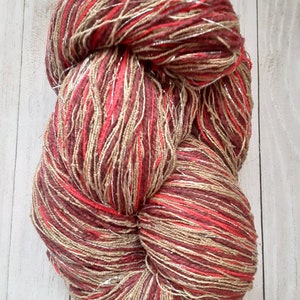 Op de afbeelding: Een bol garen met een mix van rode, bruine en beige kleuren. Het garen heeft een metallic zilveren draad die erdoorheen is geweven.