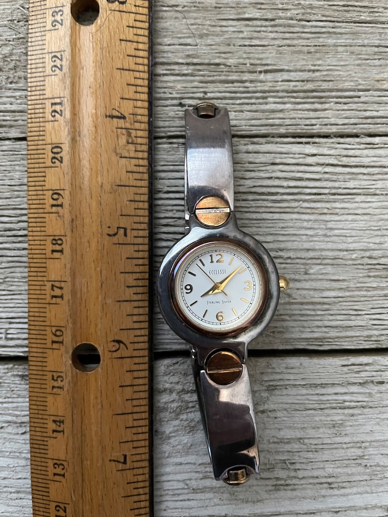 May include: A silver and gold tone wristwatch with a white face and black hands. The watch face has the text "ECCLISSI STERLING SILVER" on it.