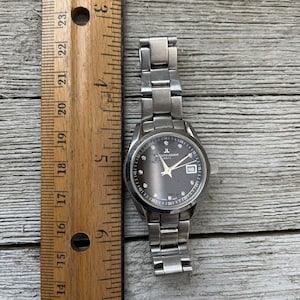 May include: A silver wristwatch with a black face and silver hands. The watch has a silver metal band and a date window at the 3 o'clock position. The watch face has a silver ring around the edge with numbers and markings. The watch face also has a small silver ring around the center with the words "Jacques Lemans" and "Swiss Made" printed on it.