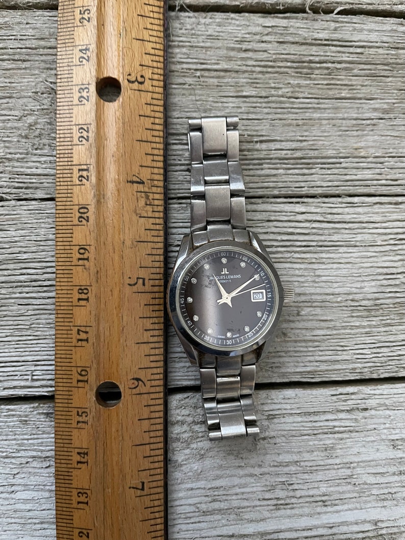 May include: A silver wristwatch with a black face and silver hands. The watch has a silver metal band and a date window at the 3 o'clock position. The watch face has a silver ring around the edge with numbers and markings. The watch face also has a small silver ring around the center with the words "Jacques Lemans" and "Swiss Made" printed on it.