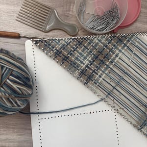 May include: A craft setup featuring a white weaving board with a partially woven blue, brown, and tan plaid pattern. A ball of yarn, a comb, a crochet hook, and a container of pins are also present, suggesting a textile project.