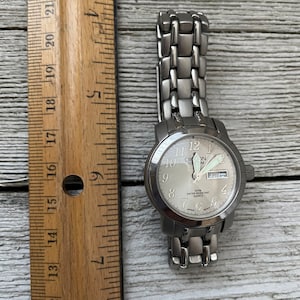 May include: A silver-toned wristwatch with a white face and black numbers. The watch has a silver metal band and says "Cron Titm" on the face. The day of the week is displayed as "FRI".