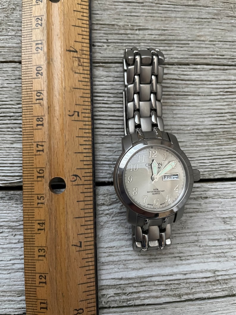 May include: A silver-toned wristwatch with a white face and black numbers. The watch has a silver metal band and says "Cron Titm" on the face. The day of the week is displayed as "FRI".