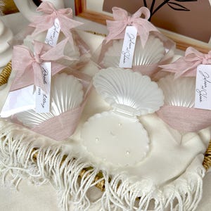 May include: White seashell-shaped candles, packaged in clear cellophane and tied with pink ribbons. Each candle has a tag that reads "Johanna & Glen". The candles are arranged in a woven basket with a white fringed cloth.