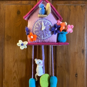 May include: A handcrafted knitted cuckoo clock in shades of lavender and pink, featuring a blue bird in the top opening, a clock face with white hands, and colourful crocheted flowers. A white mouse hangs from the base with blue weights.