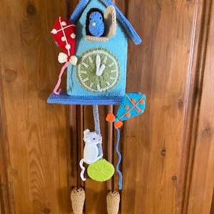 Könnte beinhalten: Eine handgestrickte Kuckucksuhr als Wandbehang in Hellblau mit grünem Zifferblatt, einem blauen Vogel im Fenster, einer weißen Maus, zwei Drachen in Rot und Blau sowie zwei beigen Gewichten, die an braunen Schnüren vor einer Holztür hängen.