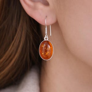 May include: Silver drop earrings with oval amber stones. The earrings have a hook closure and a silver bezel setting. The amber has a warm, golden-brown hue with visible inclusions. The earrings are a classic jewellery item.