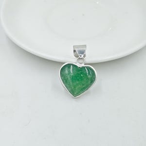 May include: A heart-shaped pendant with a vibrant green stone set in a silver-colored metal frame. The pendant is displayed on a white surface, showcasing its polished finish and unique design. A simple and elegant piece of jewelry.