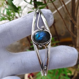 May include: A silver-toned metal cuff bracelet featuring a central oval-shaped labradorite gemstone with blue iridescence. The bracelet band has a split design with curved wire accents above and below the stone setting, creating a symmetrical look.