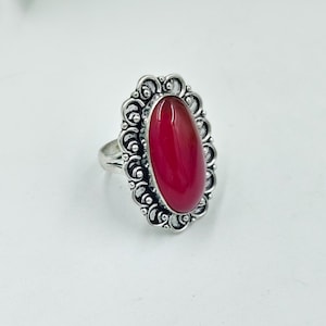 Może przedstawiać: Srebrny pierścionek z dużym, owalnym, głęboko czerwonym kamieniem szlachetnym. Kamień osadzony jest w dekoracyjnej, ażurowej, srebrnej oprawie. Obrączka pierścionka jest prosta, polerowana. Pierścionek jest prezentowany na białym tle.