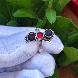 Peut inclure: Une bague en argent ornée de trois pierres : une pierre rouge centrale flanquée de deux pierres noires. La bague est présentée sur un fond de feuillage vert et une surface blanche, mettant en valeur le design et le contraste de couleurs.