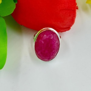 Peut inclure: Une bague en argent ornée d'une grosse pierre précieuse rose foncé de forme ovale. La pierre a une surface à facettes, captant la lumière. La bague est posée sur un fond blanc avec une fleur rouge et des feuilles vertes.