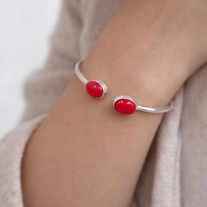 Könnte beinhalten: Ein silbernes Armreif-Armband mit zwei ovalen, leuchtend roten Steinen. Das offene Armband hat an jedem Ende einen Stein. Das Armband wird am Handgelenk getragen, mit einem neutralfarbenen Ärmel im Hintergrund.