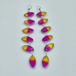 May include: A pair of long, dangling earrings. Each earring features a series of oval-shaped, gradient-coloured stones in shades of yellow and magenta, set in silver-toned metal. The earrings have a hook closure.
