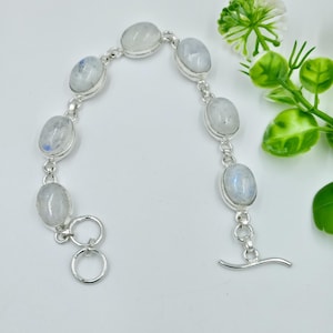 Könnte beinhalten: Ein silbernes Armband mit ovalen, irisierenden Mondstein-Cabochons. Die Steine sind mit einer Silberkette und einem Knebelverschluss verbunden. Das Armband wird auf weißem Hintergrund mit Grünzeug präsentiert.