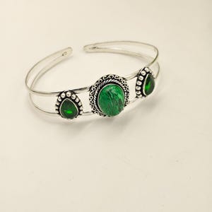 May include: Silver-toned cuff bracelet featuring three green gemstone accents. The central stone is round, surrounded by a decorative border, while the side stones are teardrop-shaped. The bracelet has a simple, open design.