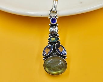 May include: Silver pendant with an oval, light green gemstone at the base, surrounded by smaller blue and yellow stones. The pendant has a loop at the top for a chain. The background is a solid yellow color.