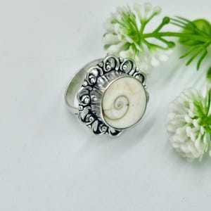 Pode incluir: Anel de prata com uma grande pedra central redonda e esbranquiçada com um desenho em espiral. O anel tem uma cravação ornamentada em estilo filigrana. Em fundo branco com flores brancas.