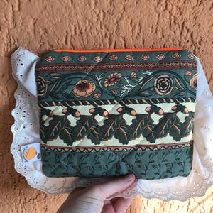 Op de afbeelding: Gewatteerde etui met een bloemen- en eikenbladpatroon in groen, perzik en crème. De etui heeft een oranje rits en wordt gepresenteerd op een wit kanten kleedje. Een klein label met een oranje afbeelding is bevestigd.