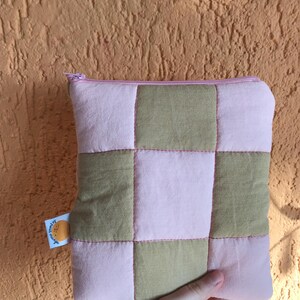 Peut inclure: Une pochette zippée en patchwork rose et vert olive. La pochette est composée de carrés de tissu cousus ensemble avec du fil rose. Une petite étiquette avec du texte et un graphisme circulaire est attachée sur le côté.