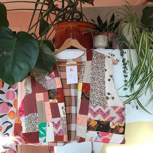 Op de afbeelding: Een patchwork vest met een verscheidenheid aan stofpatronen, waaronder bloemen, luipaardprint en geometrische ontwerpen. Het vest hangt aan een houten hanger. Het kleurenpalet omvat tinten bruin, roze en crème.