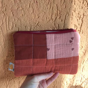 Op de afbeelding: Een gewatteerde, rechthoekige etui met een rode rits. De etui is verdeeld in vierkanten in verschillende tinten rood en roze. Kleine hartvormen zijn verspreid over de roze secties. Op een getextureerde oranje achtergrond.