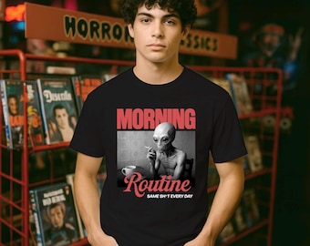 Pode incluir: Camiseta preta com um gráfico de um alienígena fumando um cigarro, uma xícara de café e as palavras "MORNING ROUTINE" em rosa. A frase "SAME SH*T EVERY DAY" está impressa abaixo da imagem do alienígena. A camiseta é usada por uma pessoa.