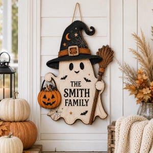 May include: A wooden wall hanging shaped like a ghost wearing a black witch hat with a crescent moon, stars, and spiderweb design. The ghost holds a broom and an orange jack-o'-lantern bucket, with the text THE SMITH FAMILY printed in black.