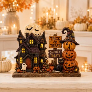 May include: A Halloween tabletop decoration featuring a dark, illuminated haunted house with a full moon, a ghost, and pumpkins. Next to it, a stack of three carved pumpkins wears a witch hat, beside a black cat and signs reading TRICK OR TREAT and SPOOKY TREATS.