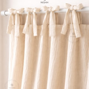 May include: Cream-colored linen curtains with tie-top closures. The curtains hang from a white rod, with each panel secured by a fabric bow. The fabric has a textured appearance. The brand name "NISHH" is visible.
