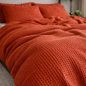 Può includere: Un letto con una coperta e cuscini color ruggine a trama a nido d'ape. La biancheria da letto è disposta su un telaio del letto con una testiera grigia. Una lampada e una piccola pianta sono visibili sullo sfondo.