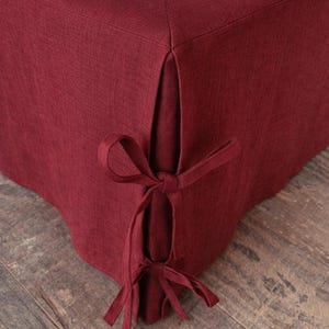 Puede incluir: Una falda de cama burdeos con un diseño a medida. La tela tiene una apariencia texturizada. La falda presenta un diseño de esquina dividida con lazos decorativos. La falda de cama se muestra en una cama con una sábana blanca y un suelo de madera.