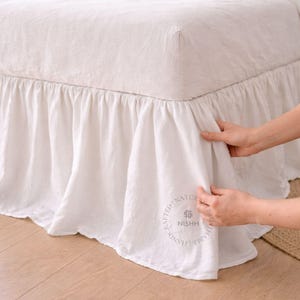 Może przedstawiać: Biała lniana spódnica do łóżka z falbanami. Spódnica do łóżka jest narzucona na łóżko z pasującą białą lnianą poszwą na kołdrę i poduszkami. Widoczne jest logo NISHH.