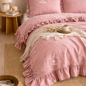 Op de afbeelding: Een roze beddengoedset met ruches, inclusief kussenslopen en een dekbedovertrek. Een kopje koffie staat op een houten dienblad. Het beddengoed ligt op een geweven vloerkleed.