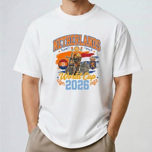 May include: A white short-sleeve t-shirt featuring a graphic print with the text NETHERLANDS World Cup 2026. The design includes a soccer ball, a windmill, a trophy, and a crest in orange, blue, and yellow colors on the chest area.