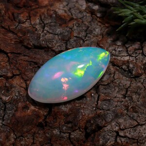 Puede incluir: Una piedra preciosa de ópalo en cabujón de forma ovalada con una base blanca translúcida y destellos vibrantes de verde neón, azul brillante y rosa suave que juegan sobre su superficie lisa, pulida y abovedada, mostrando un juego de color iridiscente.