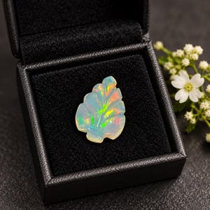 May include: A carved opal gemstone shaped like a leaf, displaying iridescent flashes of green, orange, and yellow. The translucent stone rests inside a square black jewelry box with a soft, dark interior lining, showcasing its play of color.