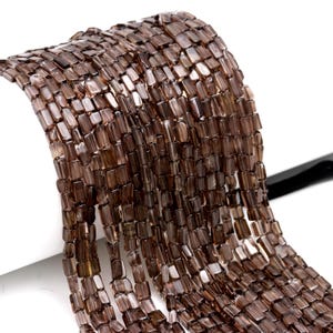 Puede incluir: Primer plano de hilos de cuentas rectangulares marrones. Las cuentas están ensartadas, creando una apariencia fluida y texturizada. Las cuentas tienen una calidad translúcida, permitiendo el paso de la luz. El fondo es blanco, resaltando las cuentas.