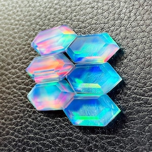 Peut inclure: Six pierres précieuses hexagonales iridescentes avec une finition polie. Les gemmes affichent une gamme de couleurs, dont le bleu, le rose et le vert, avec un effet opalescent chatoyant. Les pierres sont disposées sur une surface noire texturée.