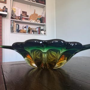 May include: A decorative glass bowl with a unique shape and color gradient. The bowl transitions from a dark green at the top to a vibrant yellow-orange at the base. The bowl has extended, wing-like edges.