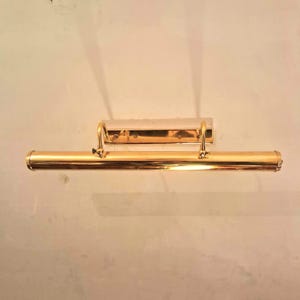 May include: A gold-toned picture light fixture with a rectangular backplate and a long, cylindrical light bar. The fixture has a classic design with curved supports and is mounted on a neutral-colored wall.