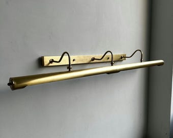 May include: A brass-toned picture light fixture. The fixture has a long, cylindrical light bar with a small, round end. It is mounted on a rectangular backplate with three curved arms. The light is designed to illuminate artwork.