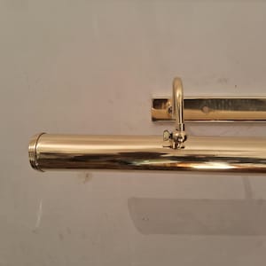 May include: A polished brass picture light fixture. The fixture has a cylindrical light housing with a curved arm and a rectangular mounting plate. The brass finish reflects the light, creating a warm glow.