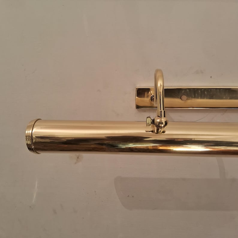 May include: A polished brass picture light fixture. The fixture has a cylindrical light housing with a curved arm and a rectangular mounting plate. The brass finish reflects the light, creating a warm glow.