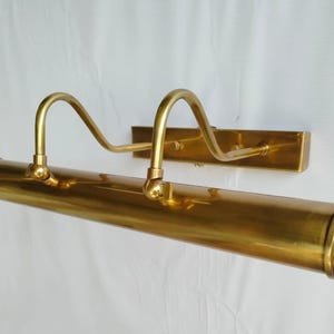 May include: A brass picture light fixture with a rectangular wall mount and a cylindrical light housing. Two curved arms extend from the housing. The fixture is a warm gold color.